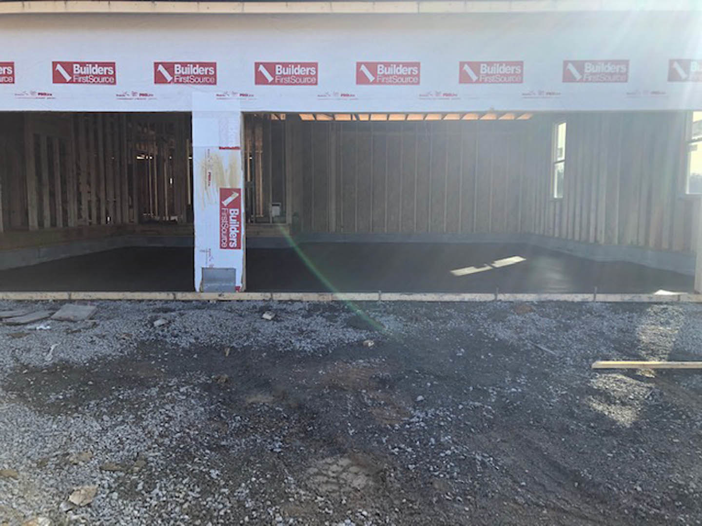 Partially built residential structure with exposed concrete walls, dirt and rocks covering the ground, white sign with red text mounted on exterior, window emitting light from