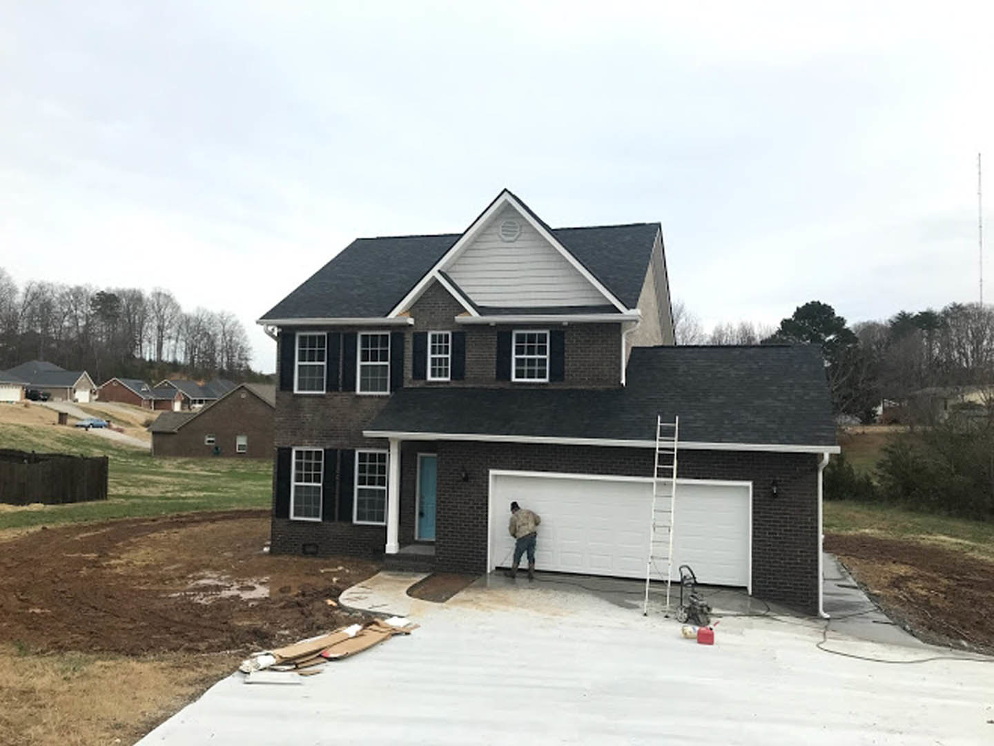 Man in white coat standing beside ladder near garage door, white-trimmed windows, gray roof, surrounded by trees on hillside