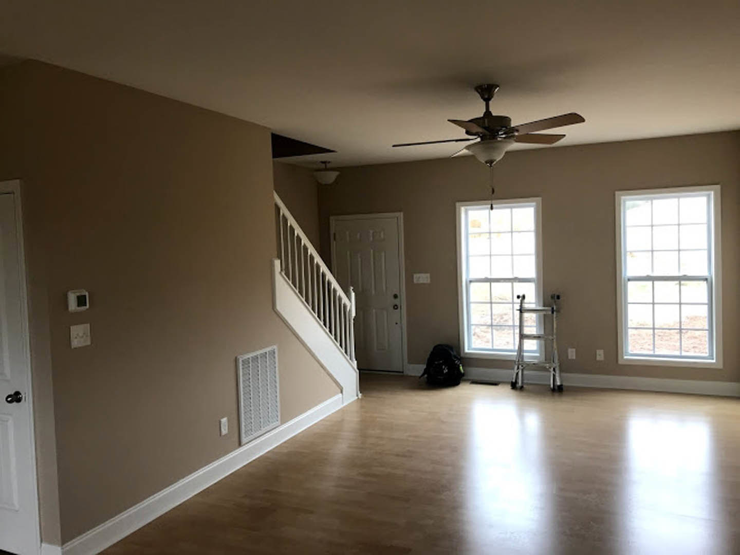 Open living area with laminate flooring, white walls, ceiling fan, staircase with wooden steps, white-framed window, and visible electrical outlet