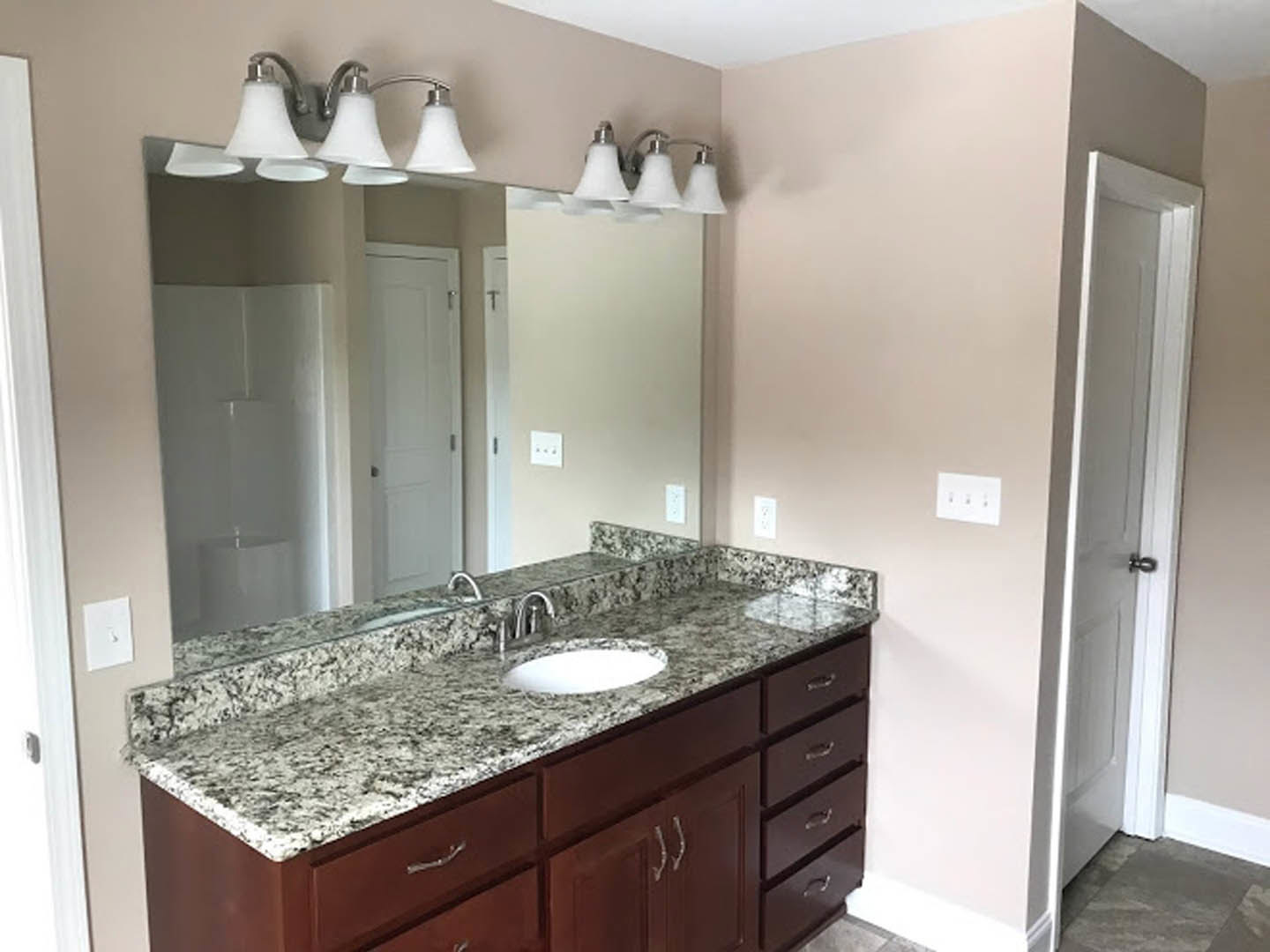 Bathroom with marble countertop, rectangular mirror above, white wall with triple light switches, black and white tile backsplash, double bulb light fixture, white cabinetry and