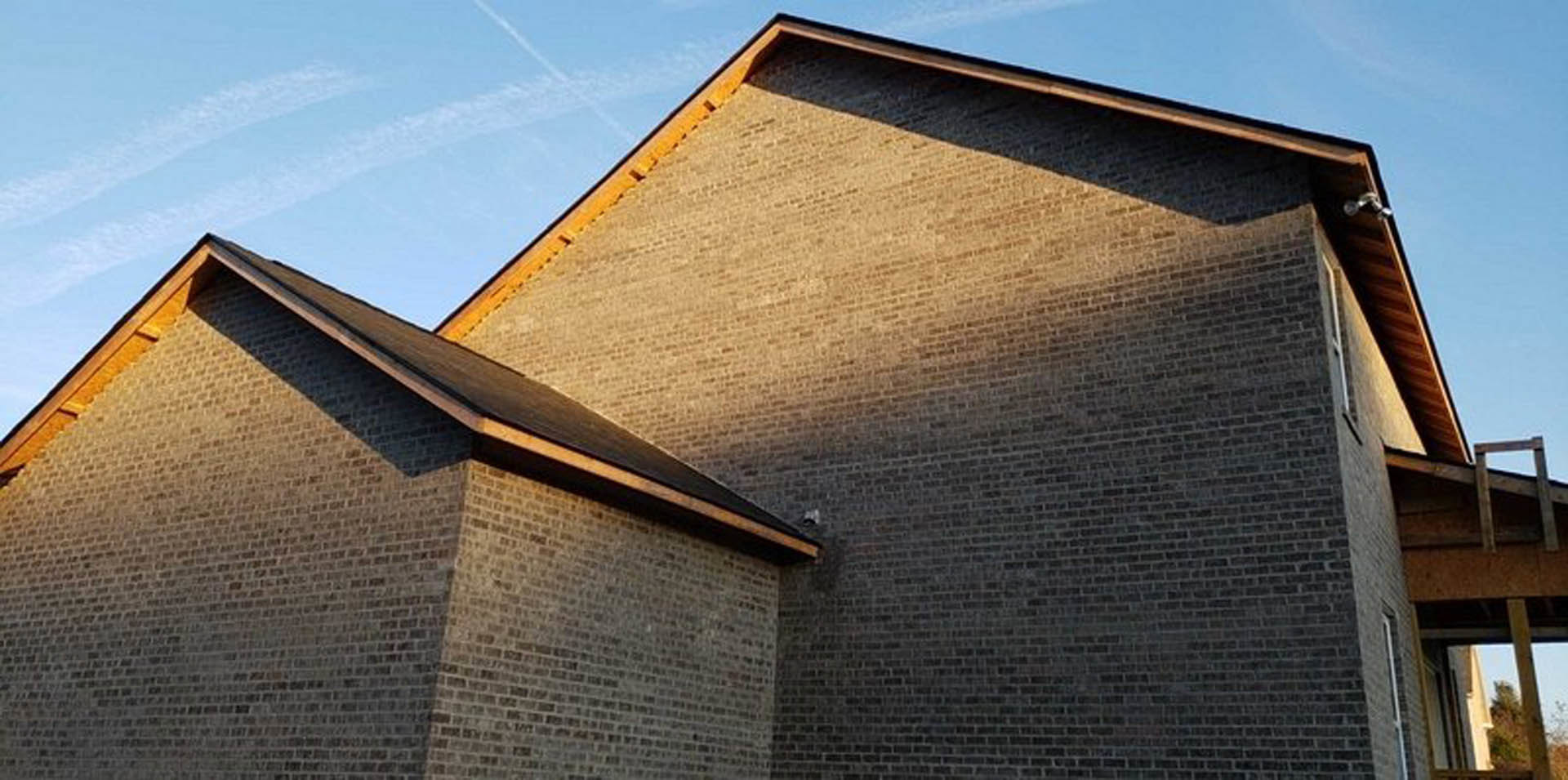 Red brick exterior wall with white-trimmed window, composite roof, and wooden beam; blue sky and blurred tree in background