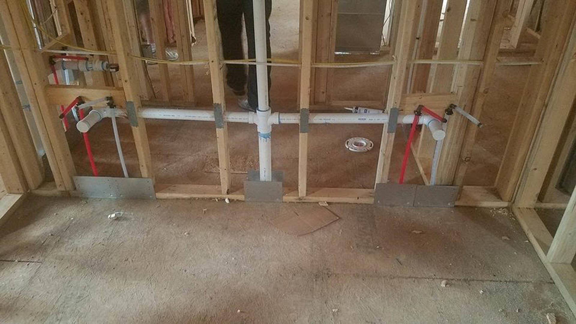 Exposed plumbing pipe set into an unfinished wall with wood framing, insulation, and construction materials visible; nearby window and floor with cardboard box.