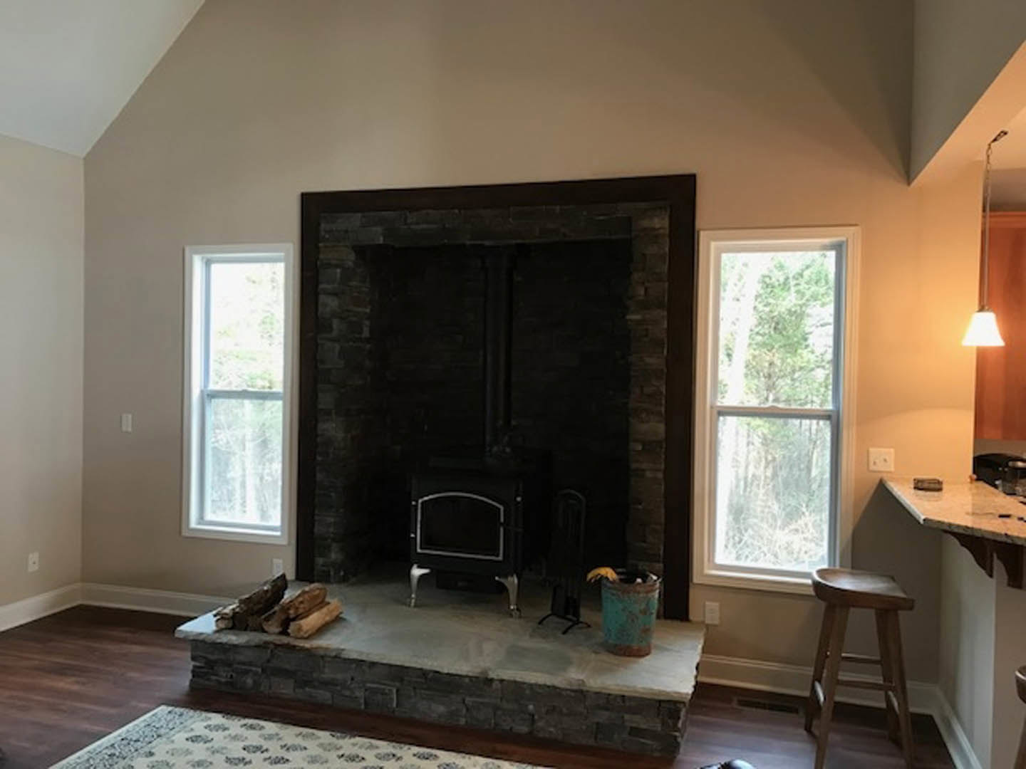 Wood-burning stove set in a fireplace with black and silver finish, wooden stool beside, green metal trash can, light fixture with fabric shade, hardwood flooring, cabinetry, and