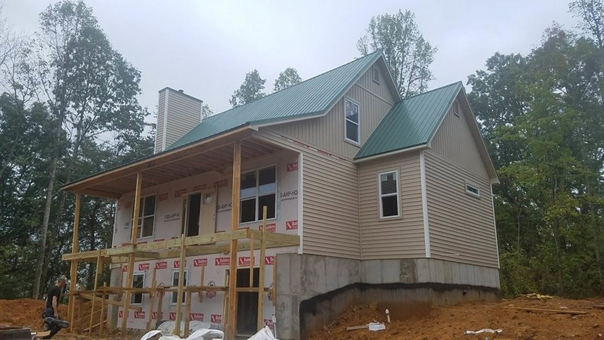 Partially built house with green roof, white-framed window, exposed framing, porch area, and water spraying from wall, surrounded by trees and sky