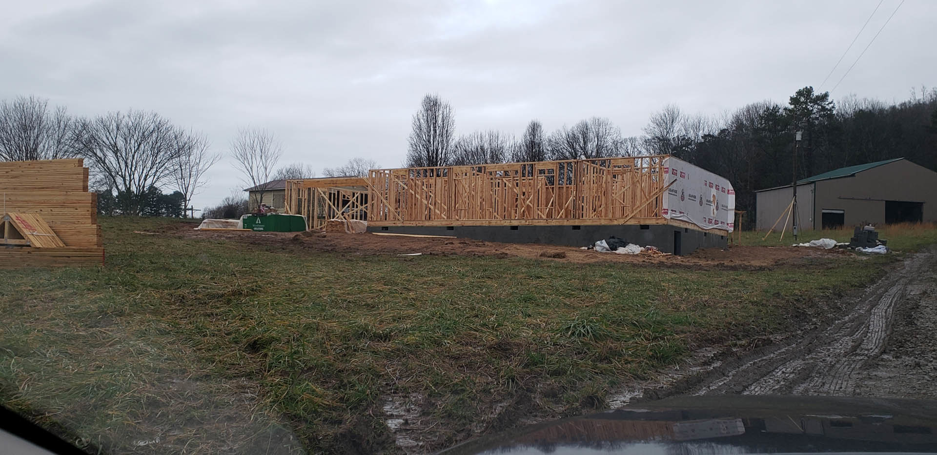 Wood-framed house under construction on grassy lot, stack of lumber in foreground, green-roofed building and trees beneath cloudy sky in background