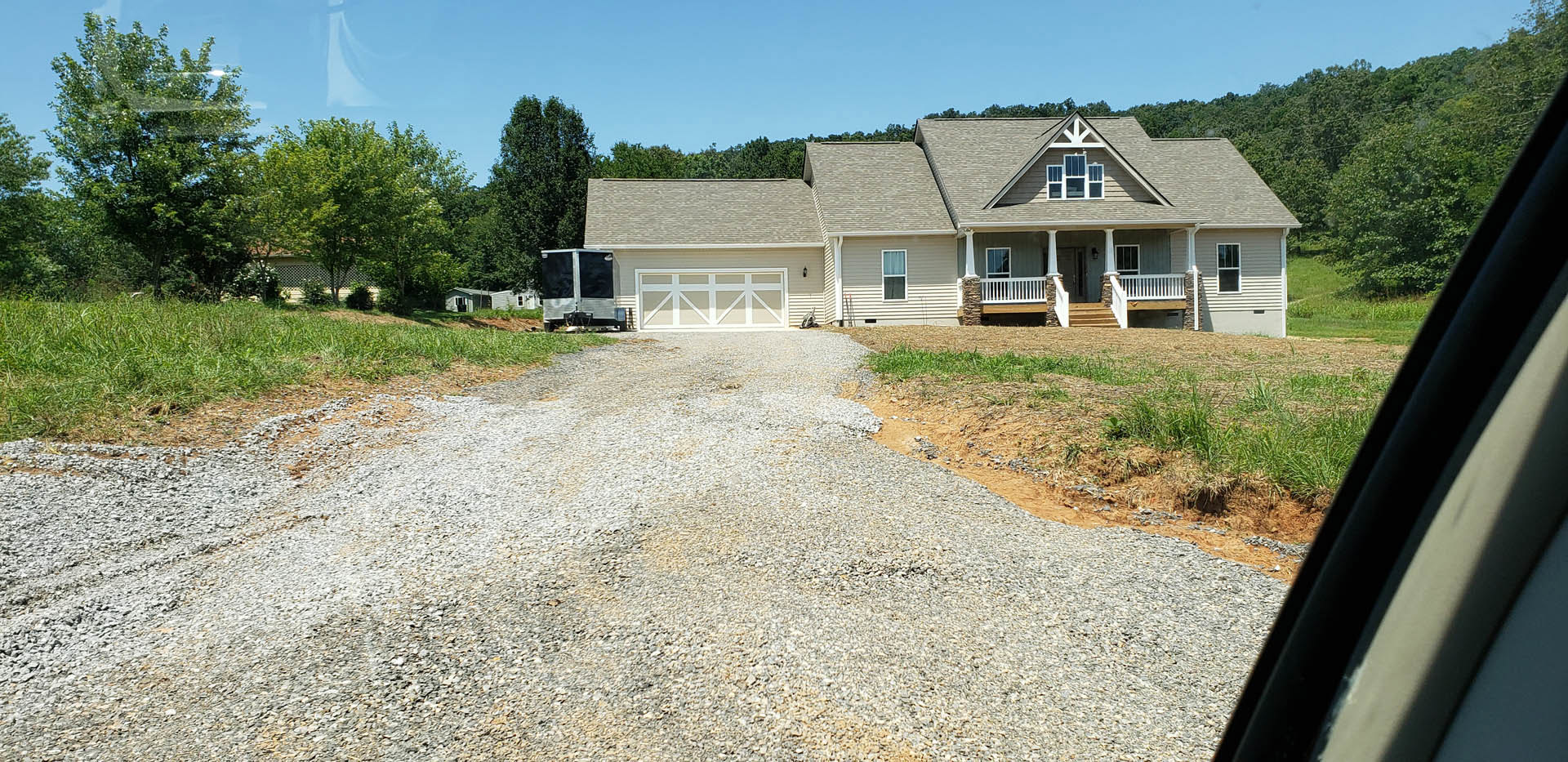 White custom home with exposed white beams, covered front porch, concrete driveway, parked trailer, grassy lawn, and mature trees under blue sky