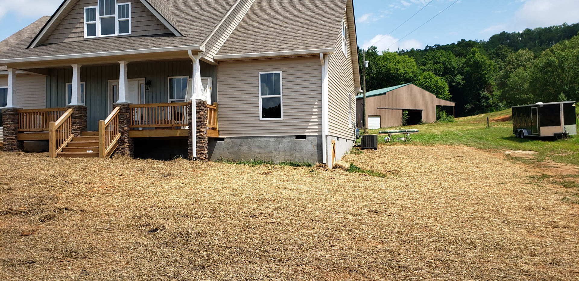 Two-story cottage with white-framed windows, white door, covered porch, grassy yard, mature trees, adjacent barn, and scattered hay on the ground