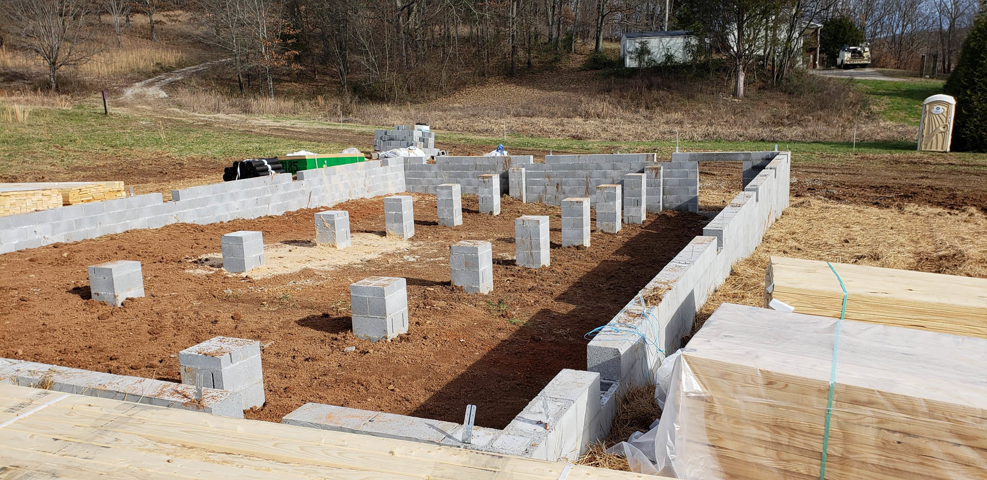Concrete foundation blocks stacked on dirt at a residential construction site, with grass and trees in the background and a portable toilet visible nearby
