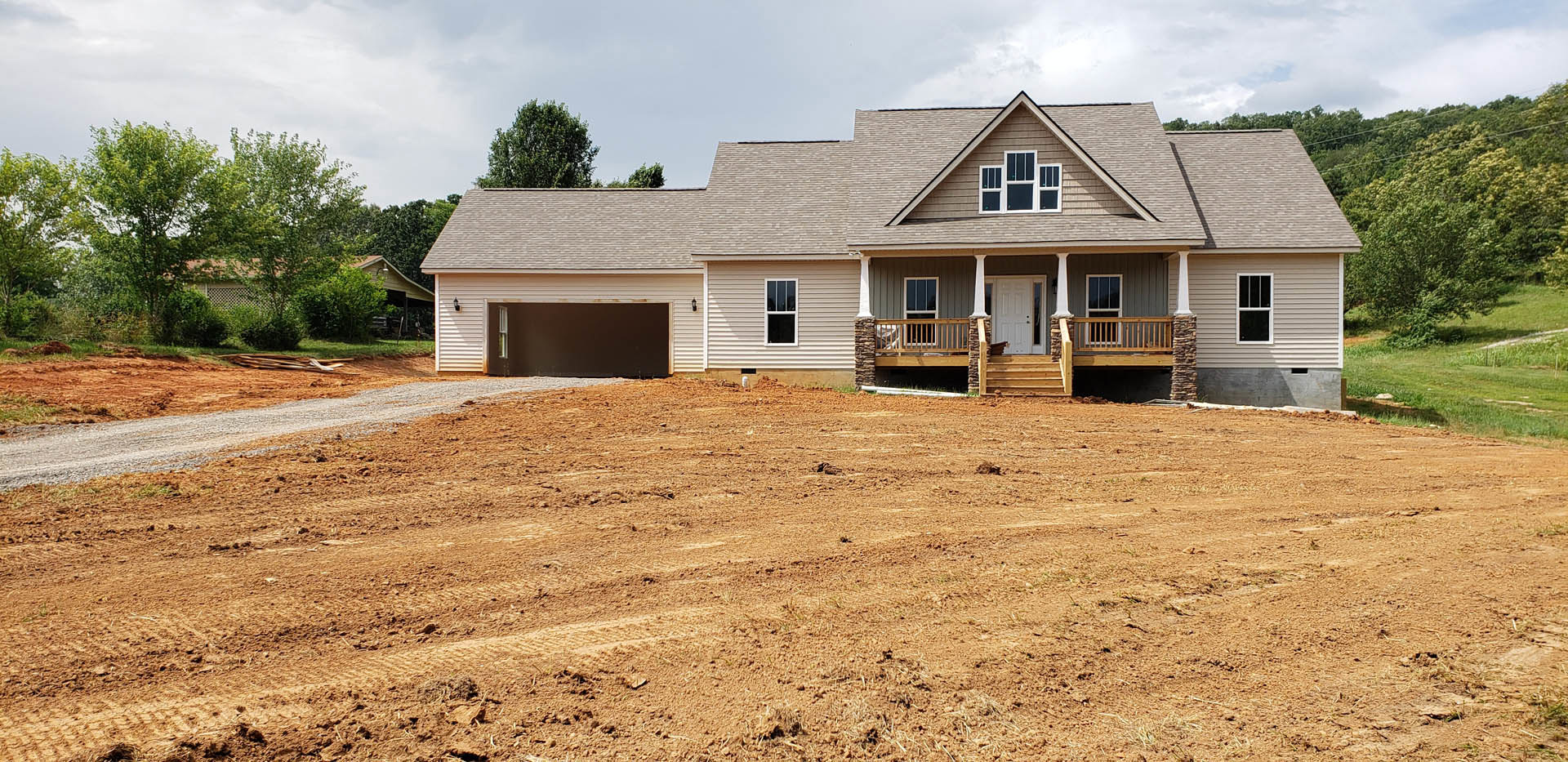 Two-story house with attached garage, white framed windows, wooden stairs leading to a white front door, large dirt field in foreground, cloudy sky above.