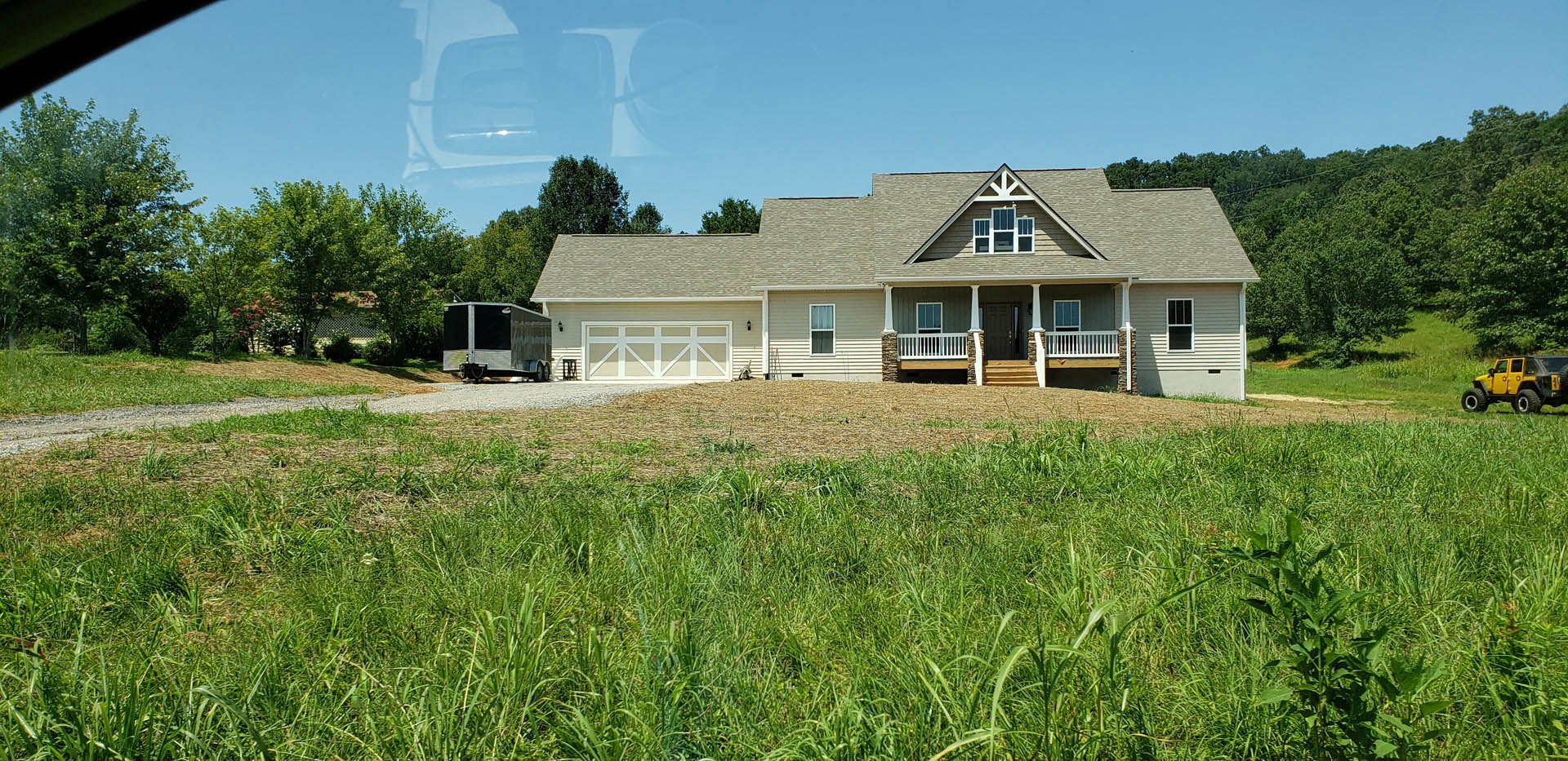 White house with covered porch and white railings, expansive green lawn in front, yellow jeep parked beside, black trailer near driveway, trees and blue sky in background.