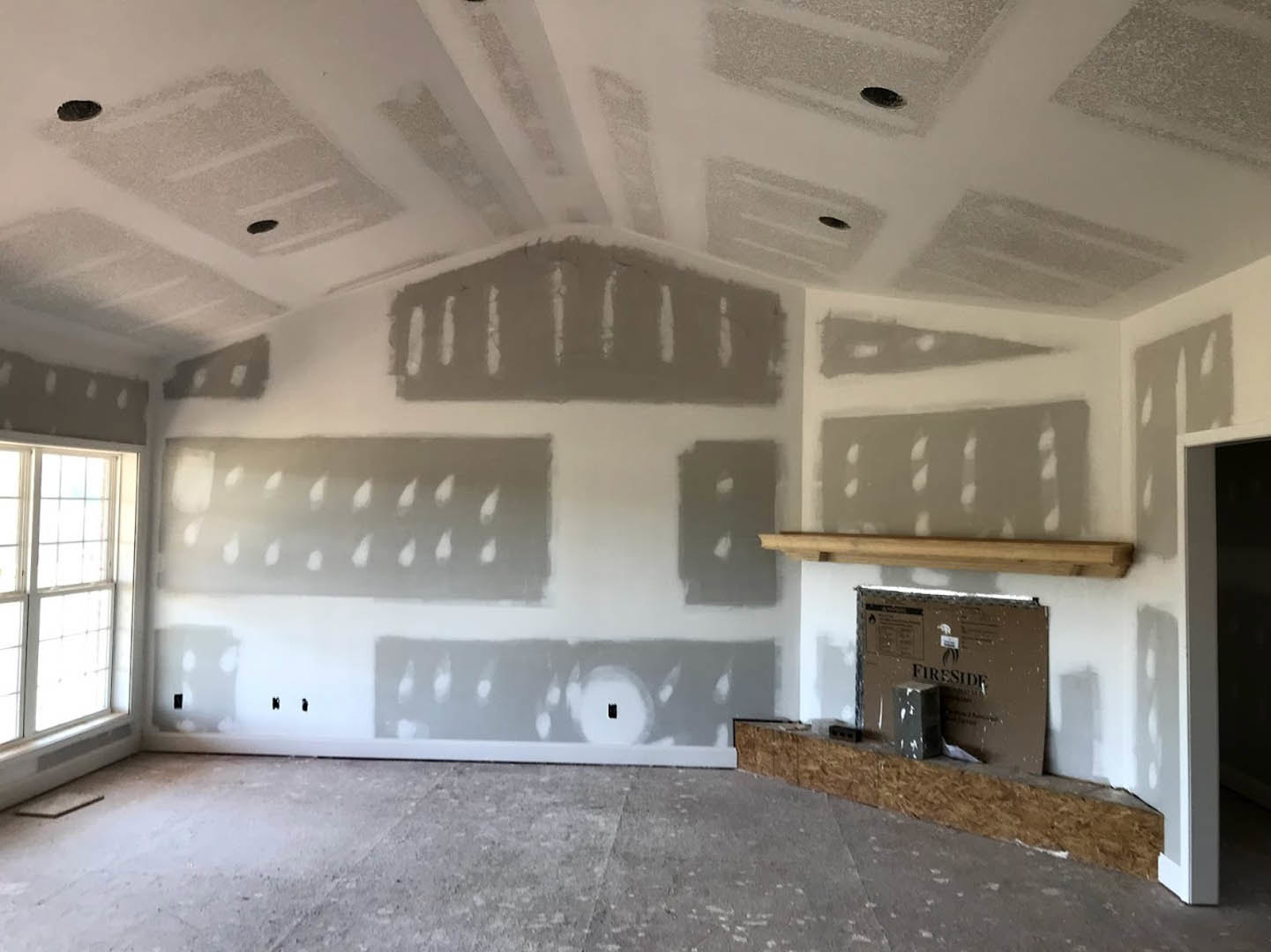 Living room with plaster walls, stone fireplace, wood flooring, and large window letting in natural light