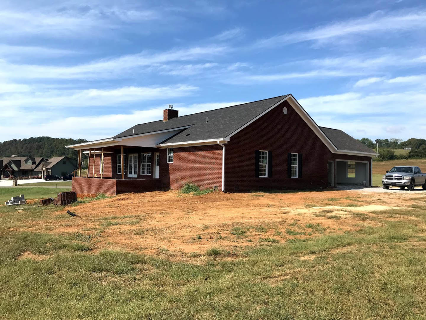 Red brick house with white roof and chimney, multi-pane windows, surrounded by green lawn under blue sky with scattered clouds, silver truck parked nearby, trees and dirt field in
