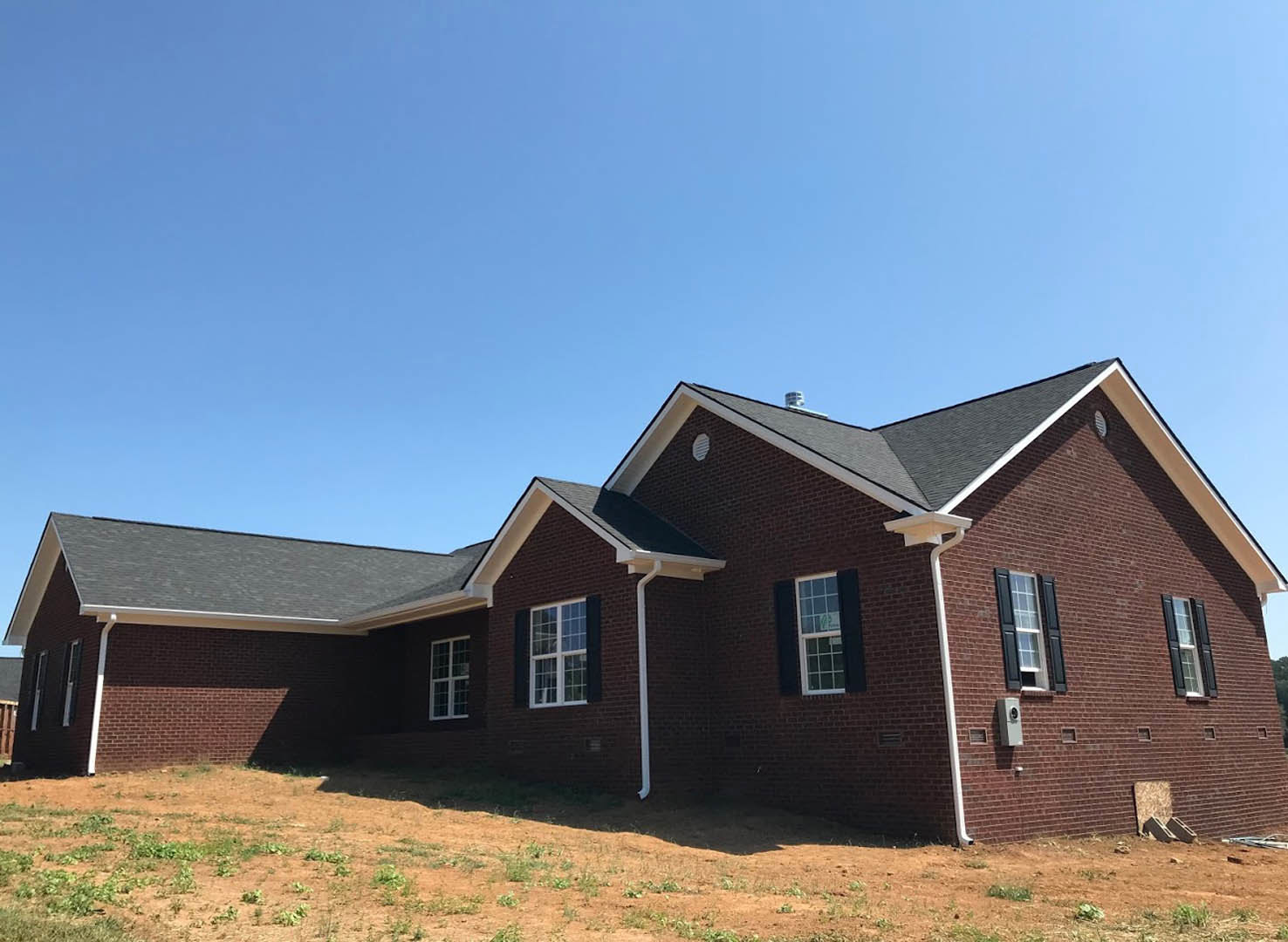 Red brick house with multi-pane window, white gutter along side, grassy yard with exposed dirt and white pipe, blue sky overhead