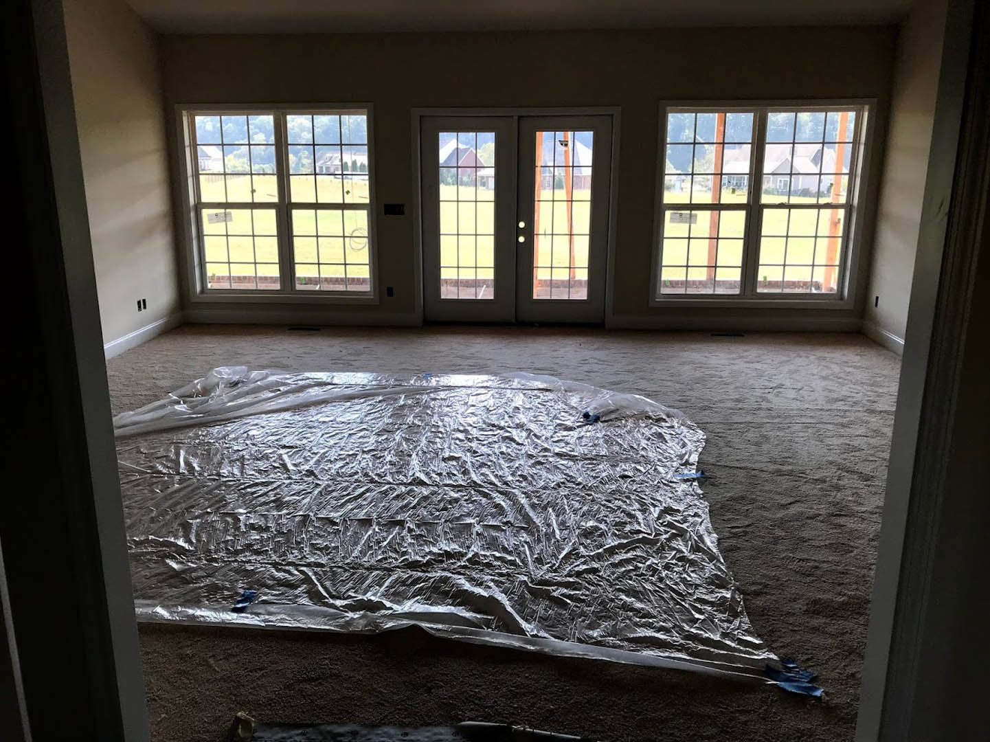 Spacious room with plastic sheet covering hardwood floor, double glass-paneled doors, large windows overlooking a field and neighboring house, white walls, abundant natural
