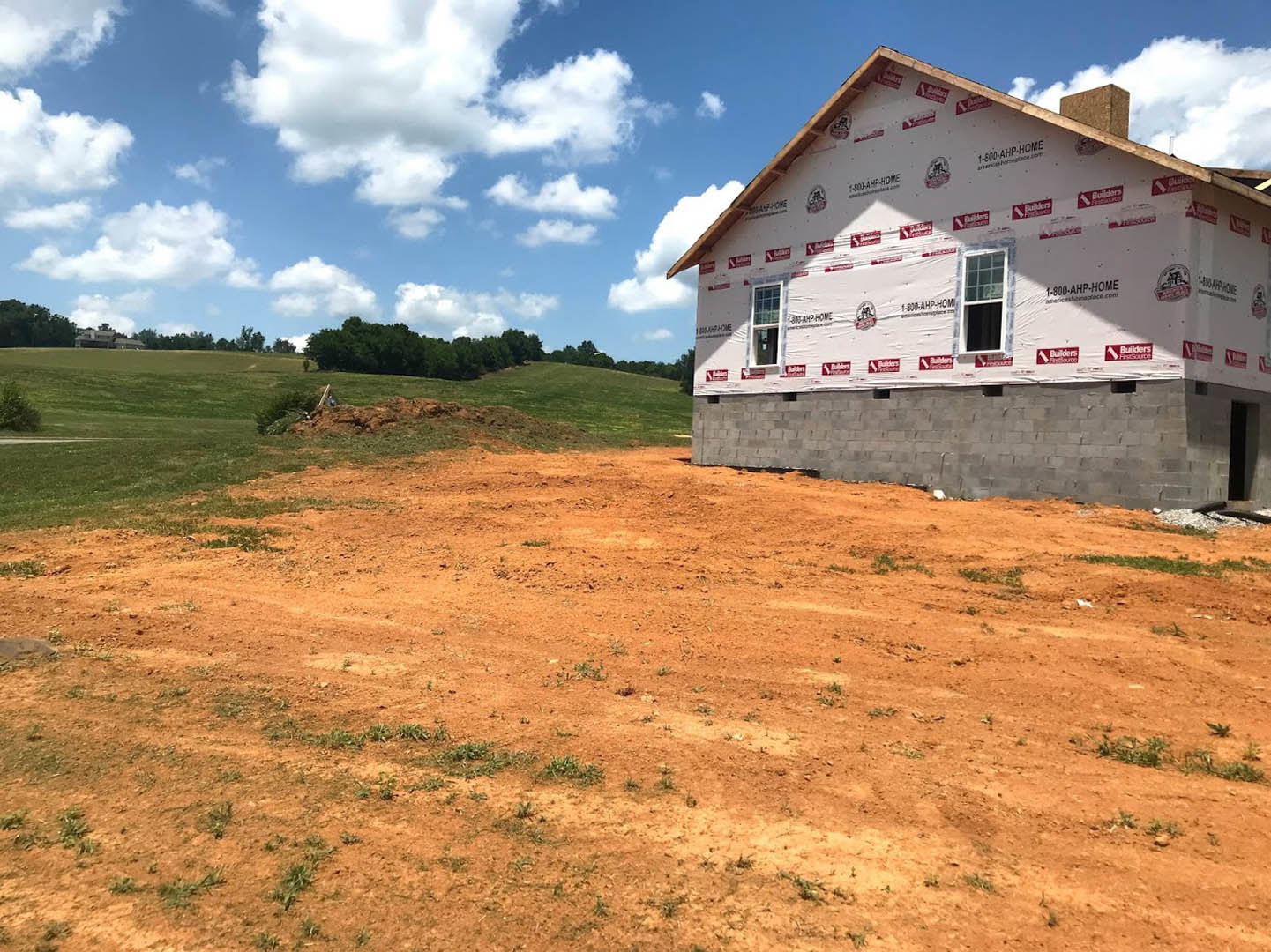 Grey brick exterior wall with white insulation sheet and red and white construction tape, multi-pane window, dirt lot, grassy rural landscape, cloudy sky.
