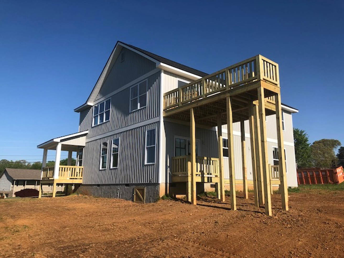 Two-story house with light siding, white-trimmed windows, covered front porch, and elevated wooden deck overlooking landscaped yard with mature trees