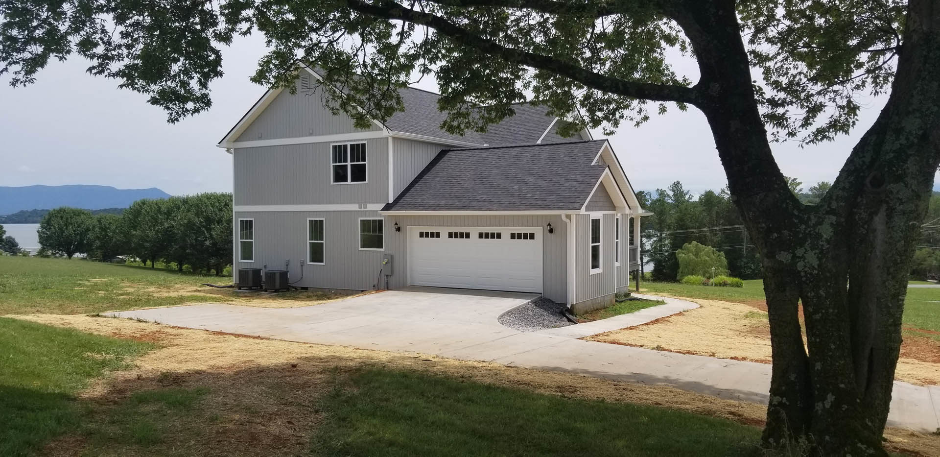 Two-story home with gray siding, attached garage, concrete driveway, large front lawn, and mature tree near the entrance.