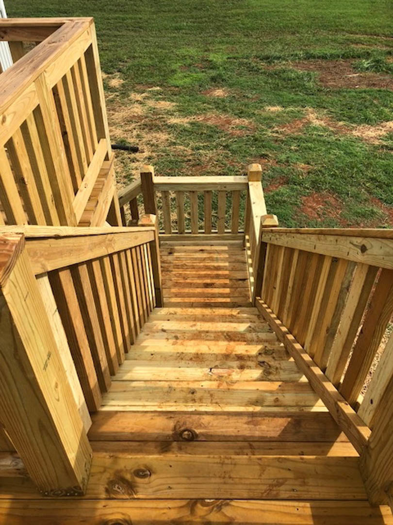 Wooden stairs with plank railings ascend from a deck to a grassy lawn, bordered by a wooden fence and handrail.