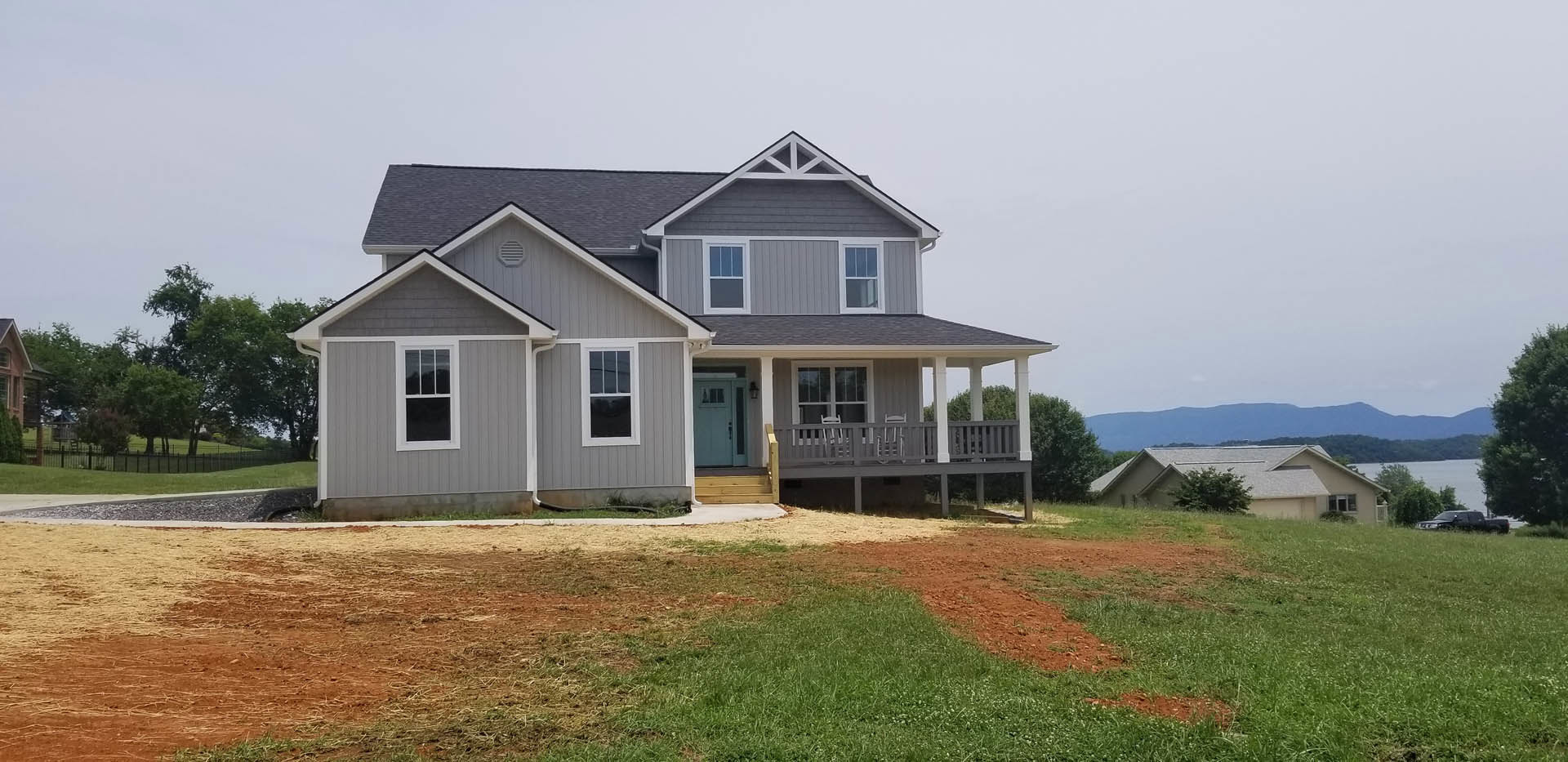 Two-story house with white-framed windows, covered porch, grassy yard, and exposed dirt area bordered by trees under a blue sky