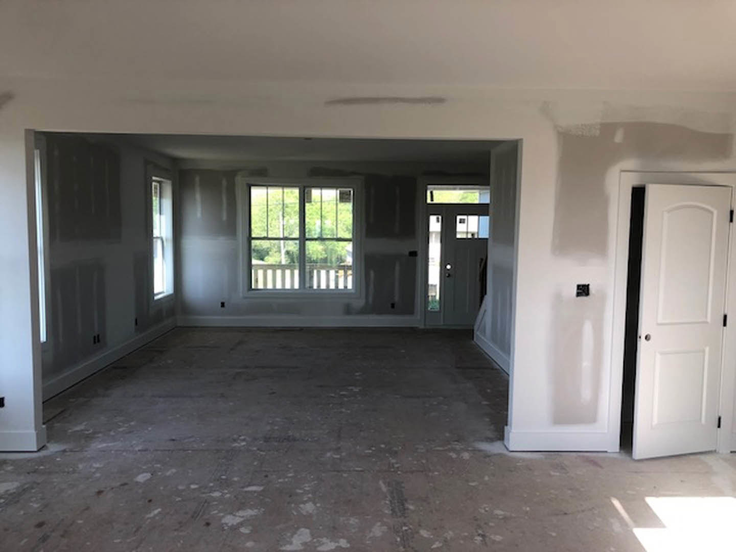 Unfinished room with white plaster walls, black-framed white door, large window overlooking trees, exposed concrete floor with white spots