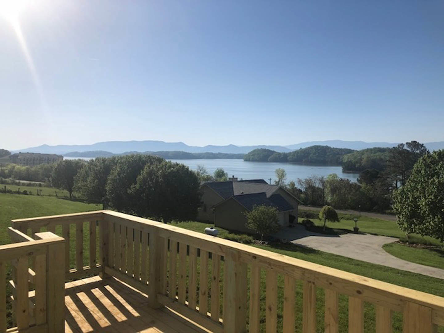 Wooden deck with railing overlooking grassy yard, lake, and distant mountains under a clear blue sky; tree branches frame the scene.