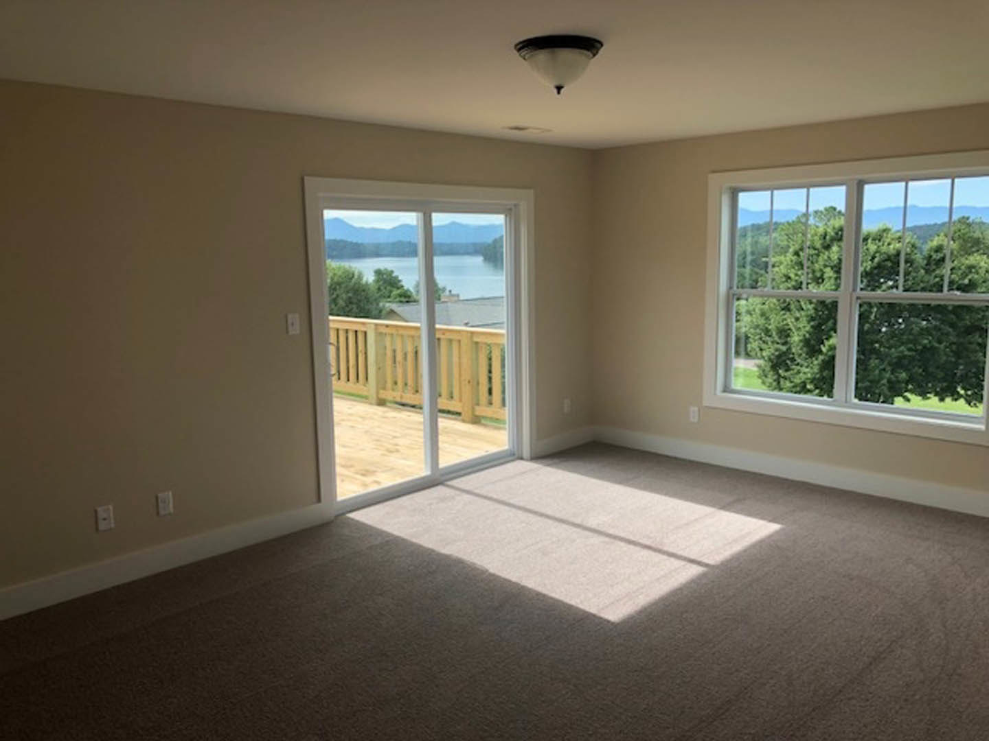 Carpeted living area with large windows and sliding glass door opening to a wooden deck, lake and trees visible outside, modern ceiling light fixture overhead