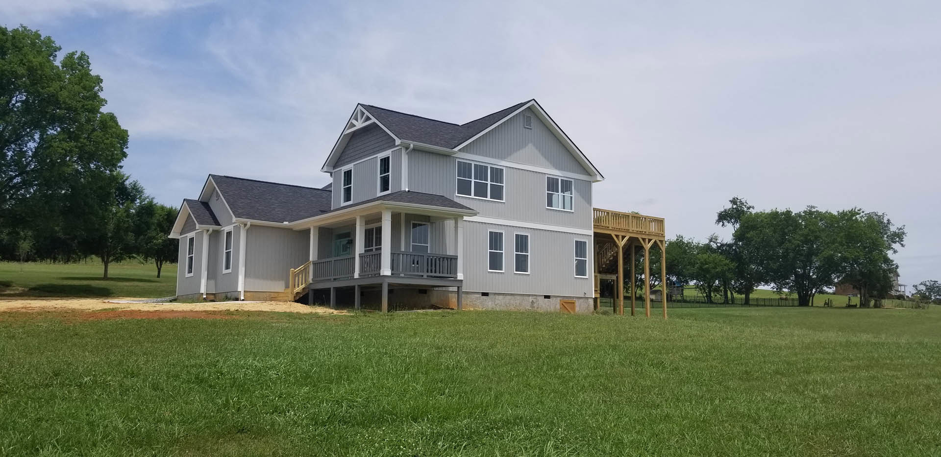 Two-story house with white siding, covered front porch, wooden deck with staircase, expansive green lawn, mature leafy tree, large windows, partly cloudy sky