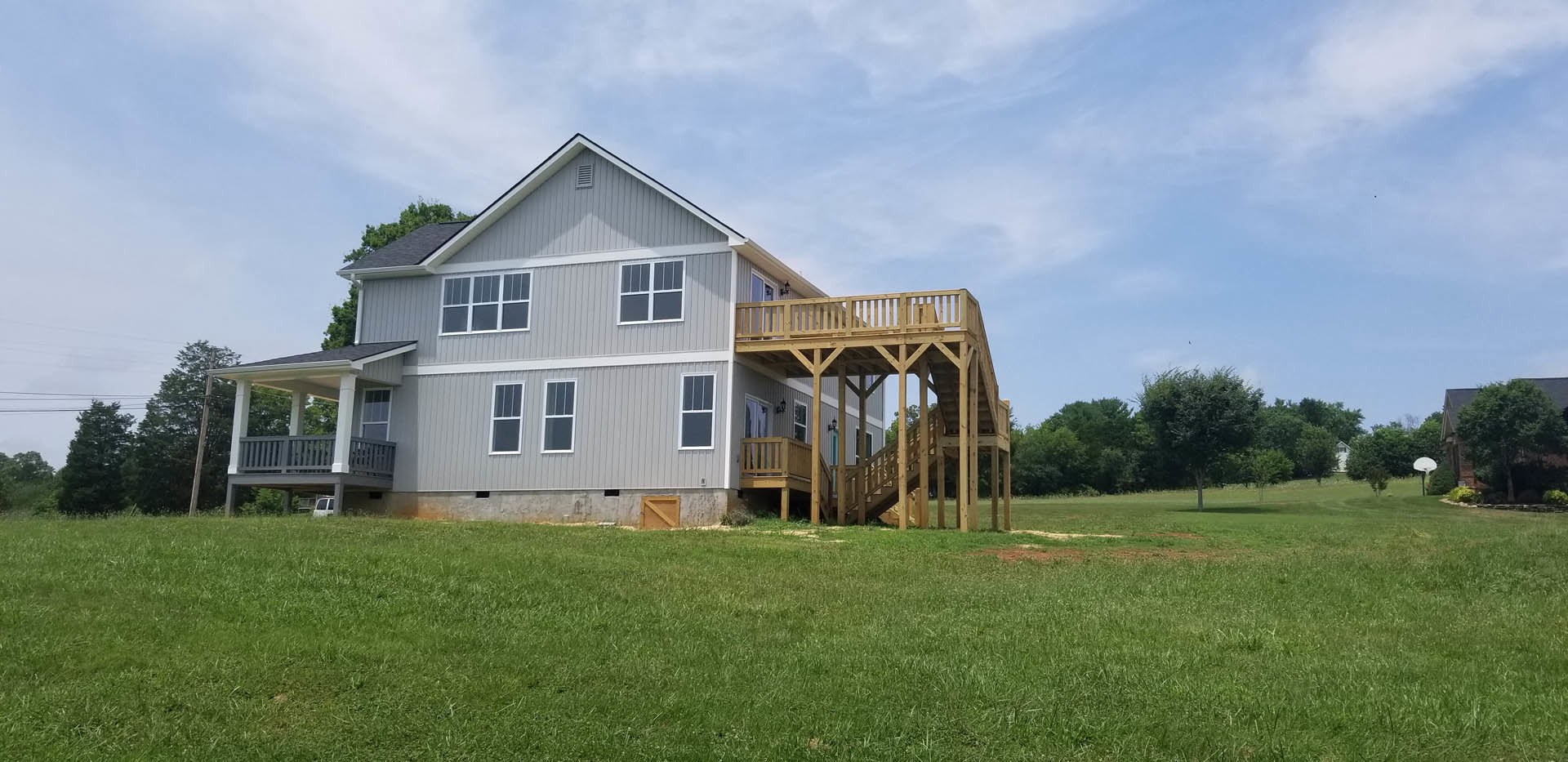 Two-story house with white siding, wooden deck and staircase, expansive green lawn, large windows with white frames, surrounded by trees under a partly cloudy sky