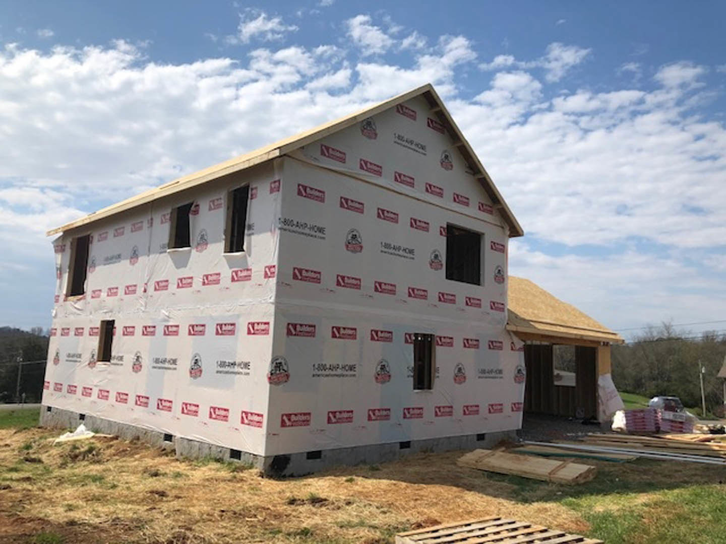 Wood-framed house under construction with exposed beams and plywood sheathing, surrounded by grass and trees beneath a clear blue sky