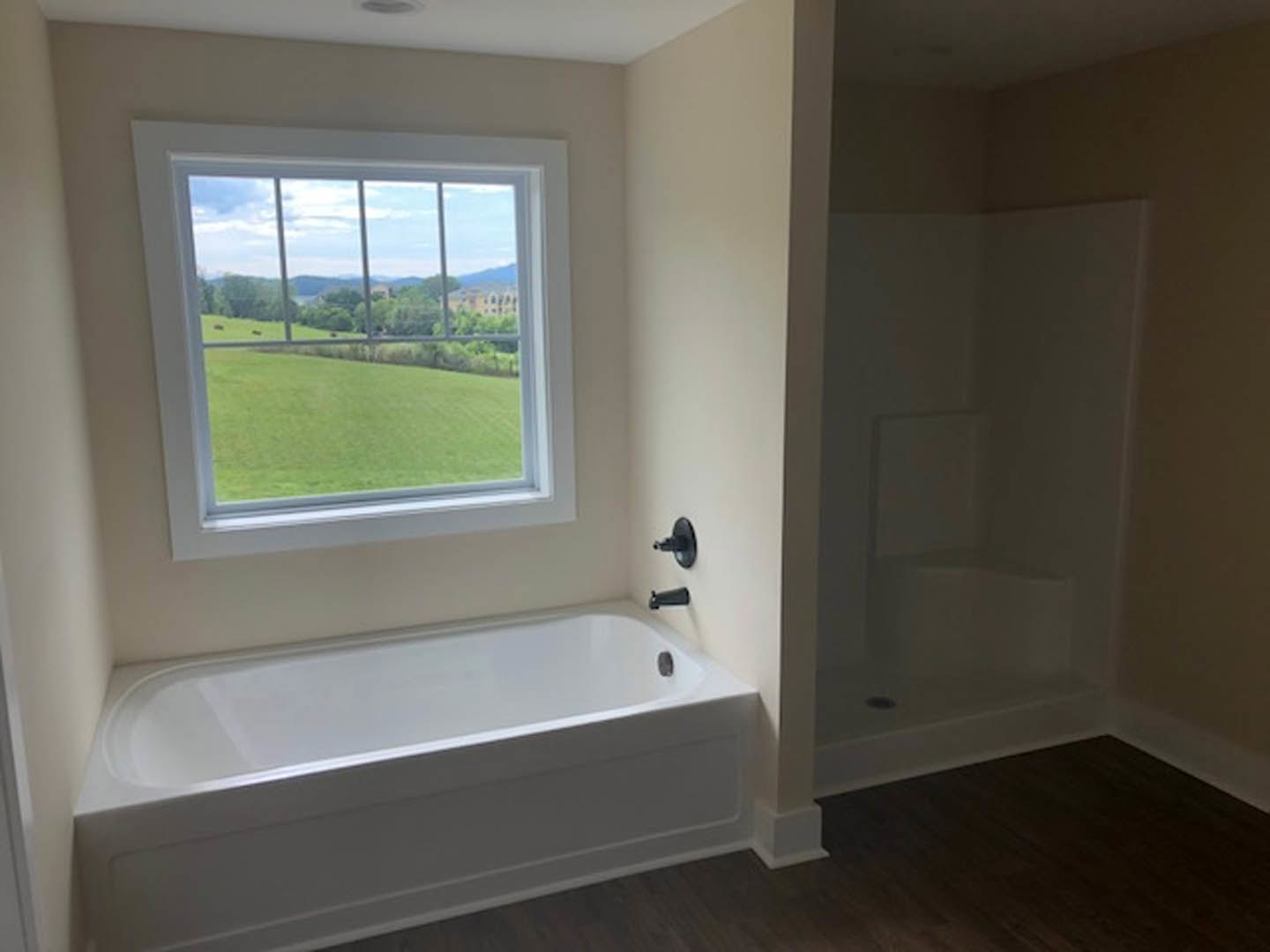 White freestanding bathtub on light wooden floor, large window overlooking grassy field, black hook mounted on white wall