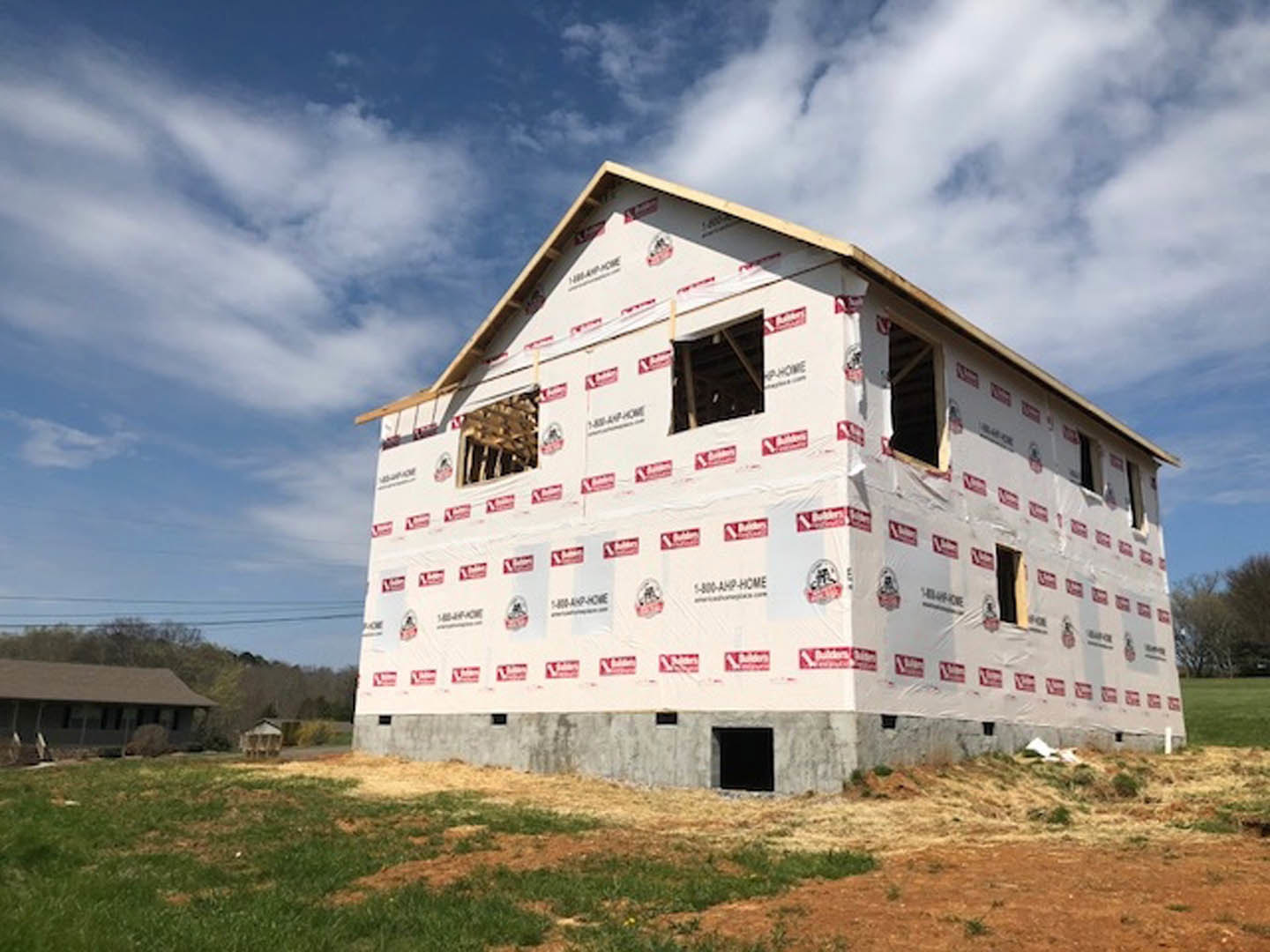 Framed custom home under construction with exposed wood, unfinished windows, and roof, set against blue sky and grassy lot with scattered trees