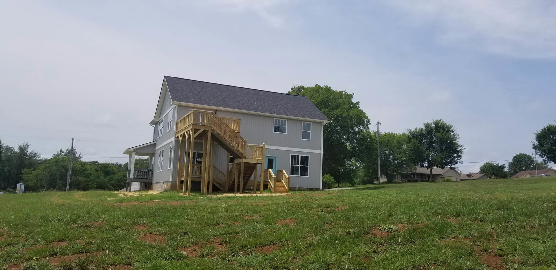 Two-story farmhouse with white siding, wooden staircase leading to elevated wooden deck, grassy yard, white-framed windows, gabled roof, and surrounding trees under partly cloudy