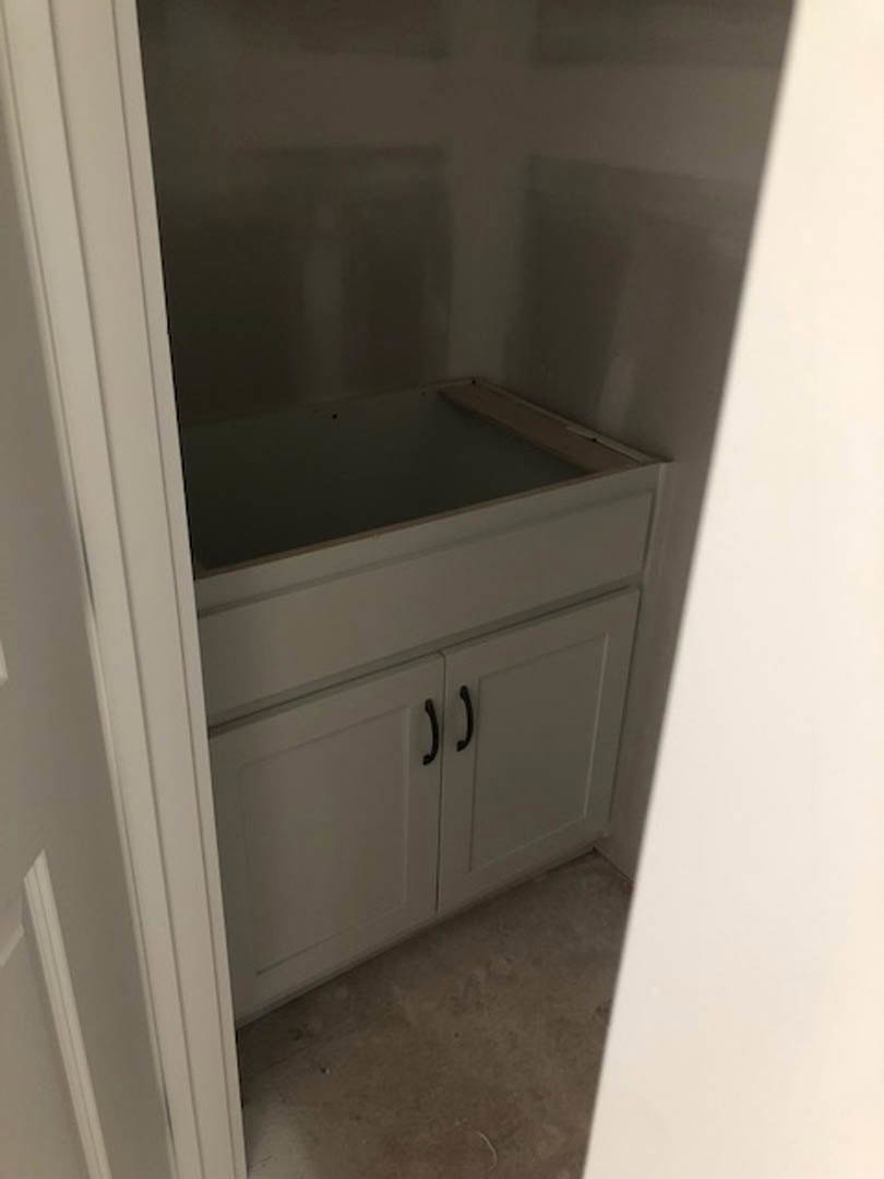 White built-in cabinet with black handles beside a white wall, shelf above, light wood floor visible below.