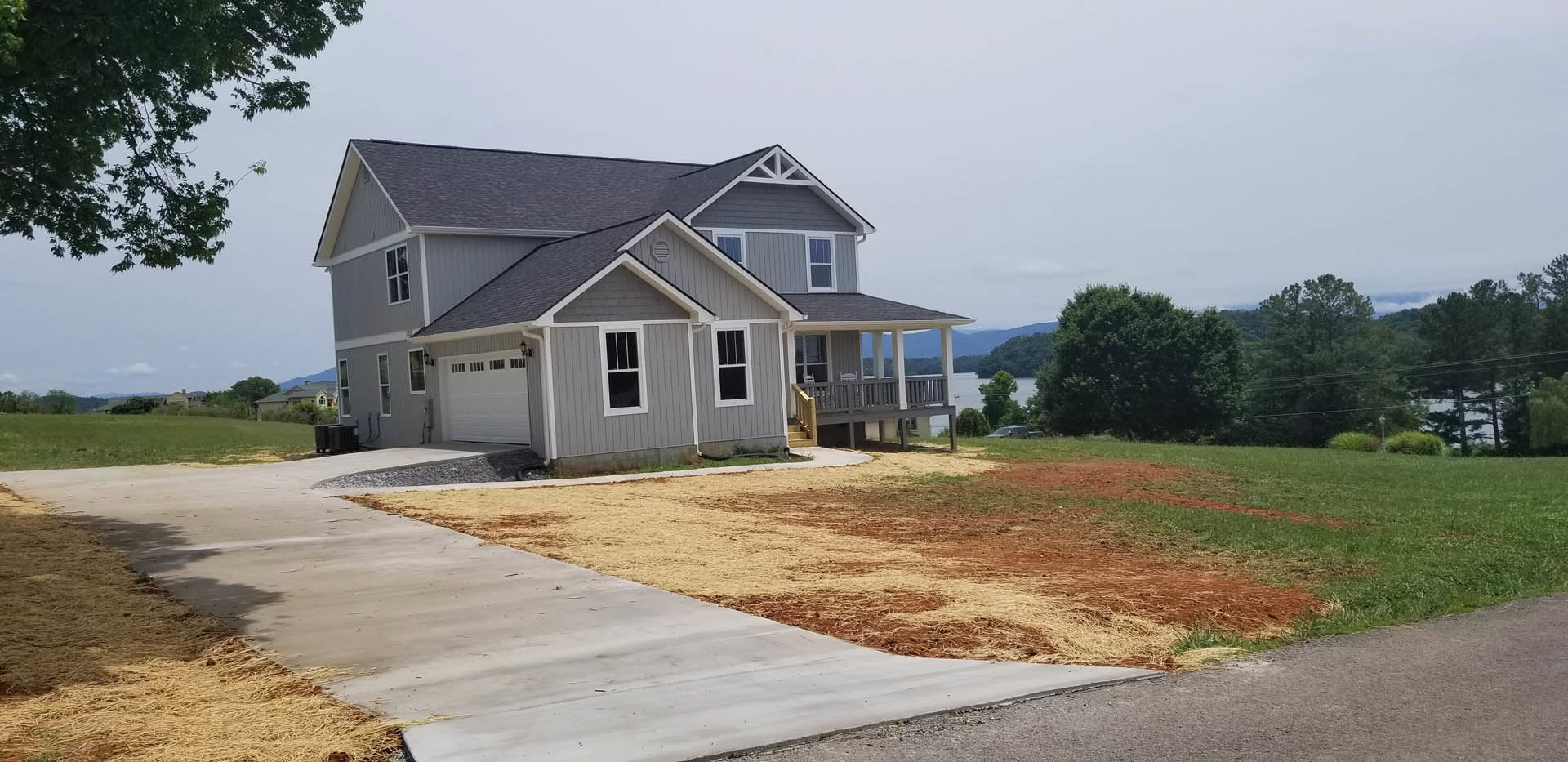 Two-story home with white-framed windows, attached garage, concrete driveway, expansive green lawn, front porch with a chair, and mature trees in the yard.