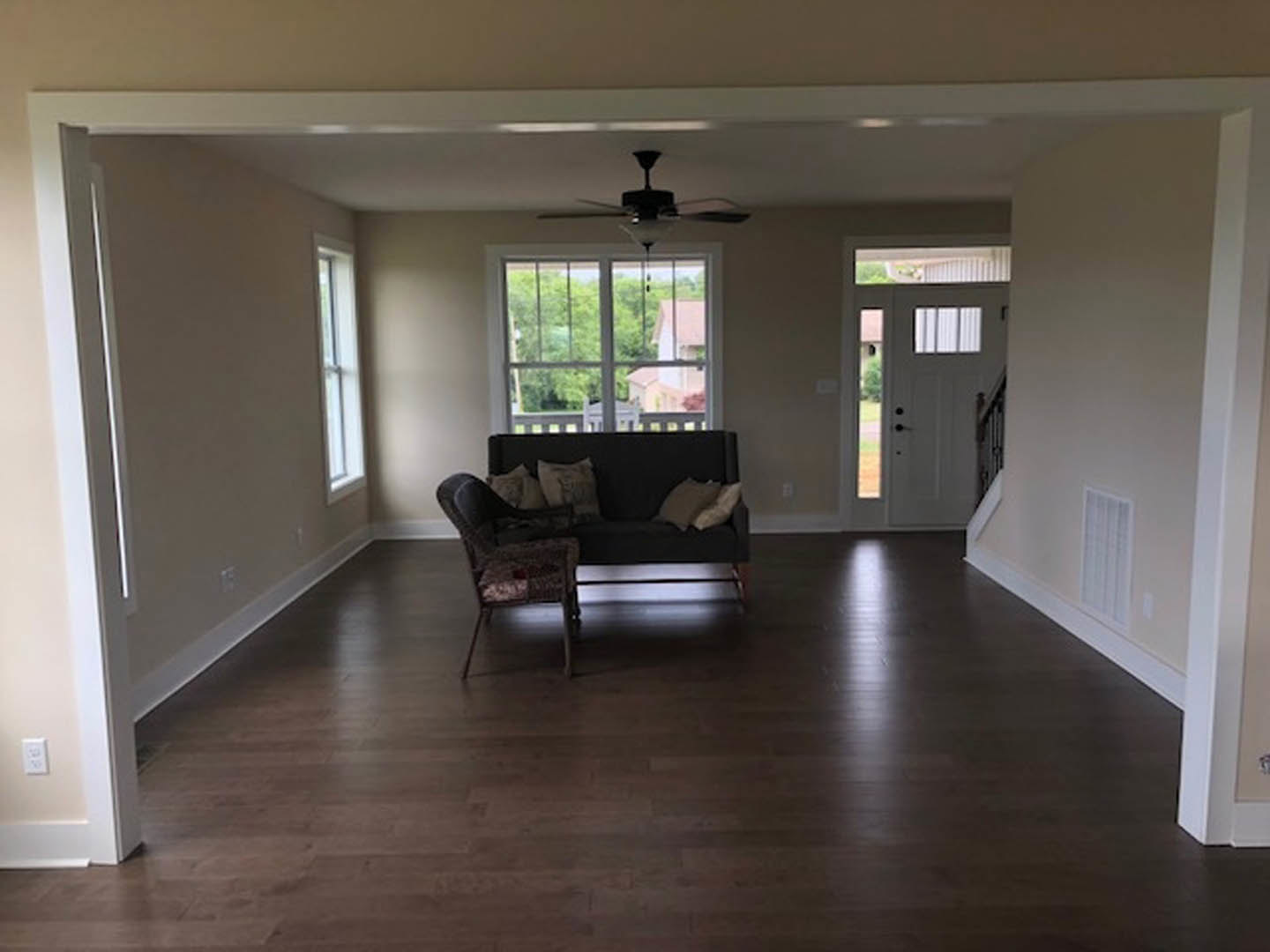Gray upholstered couch with patterned pillows in a living room featuring wood laminate flooring, white walls, a ceiling fan with light, and a window beside a white door with glass
