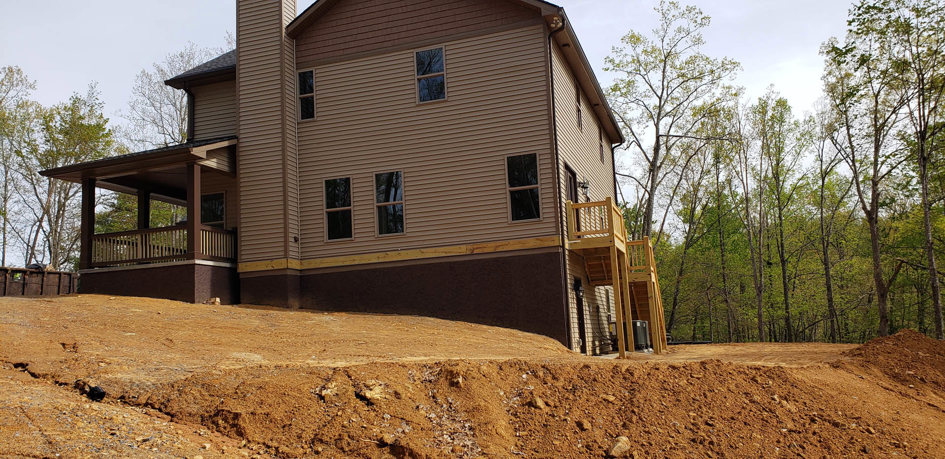 Wooden deck with railing attached to a house, dirt hill in foreground, large windows reflecting nearby trees, rural landscape with scattered plants and open sky