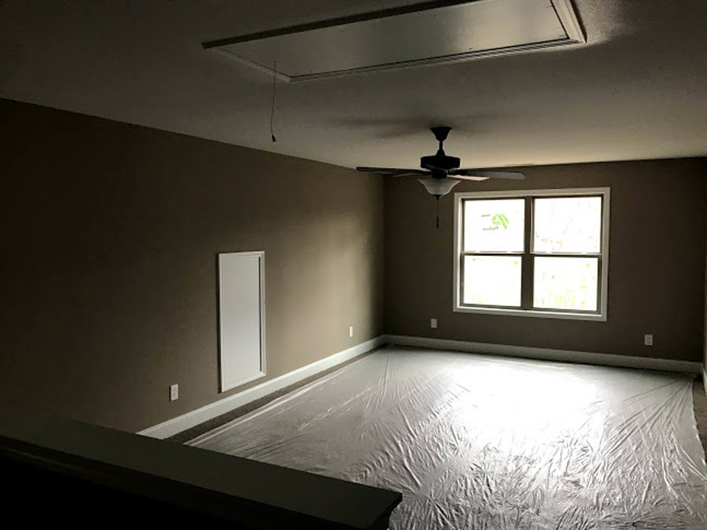 Bedroom with white plaster walls, ceiling fan with light fixture, large window featuring a logo, and white plastic covering protecting the floor.