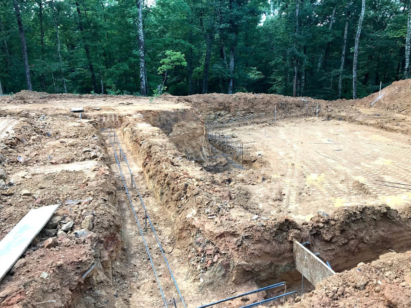 Foundation trench with exposed metal rebar, white board resting on rocky soil, pipe partially buried, dense forest of trees in background
