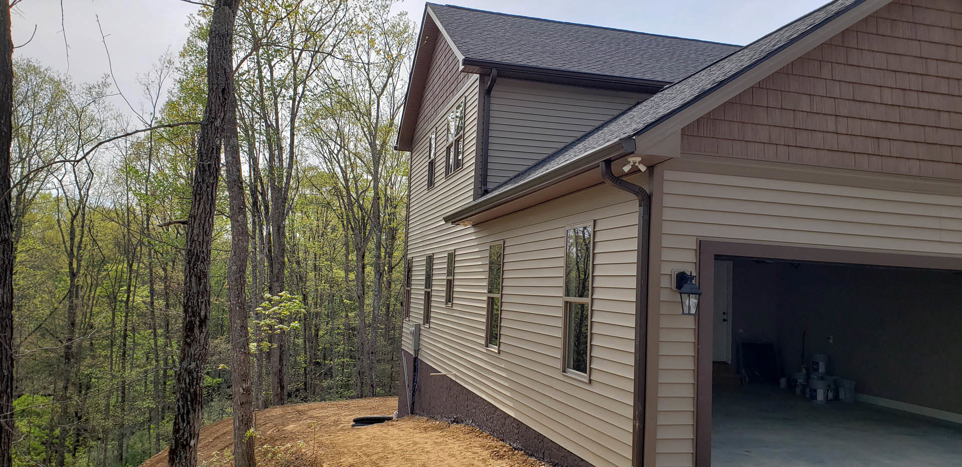 Two-story home with gray siding, large windows reflecting nearby trees, attached garage, and mature trees in the background