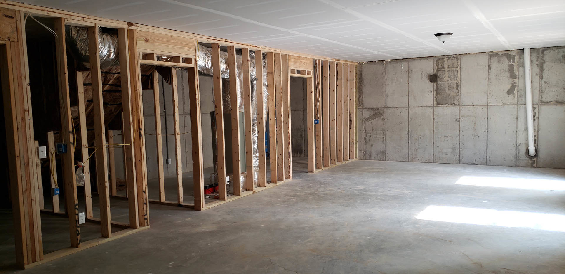 Concrete floor with exposed wood beams, wood-framed walls, white ceiling with linear detailing, white pipe mounted on wall, unfinished basement interior.