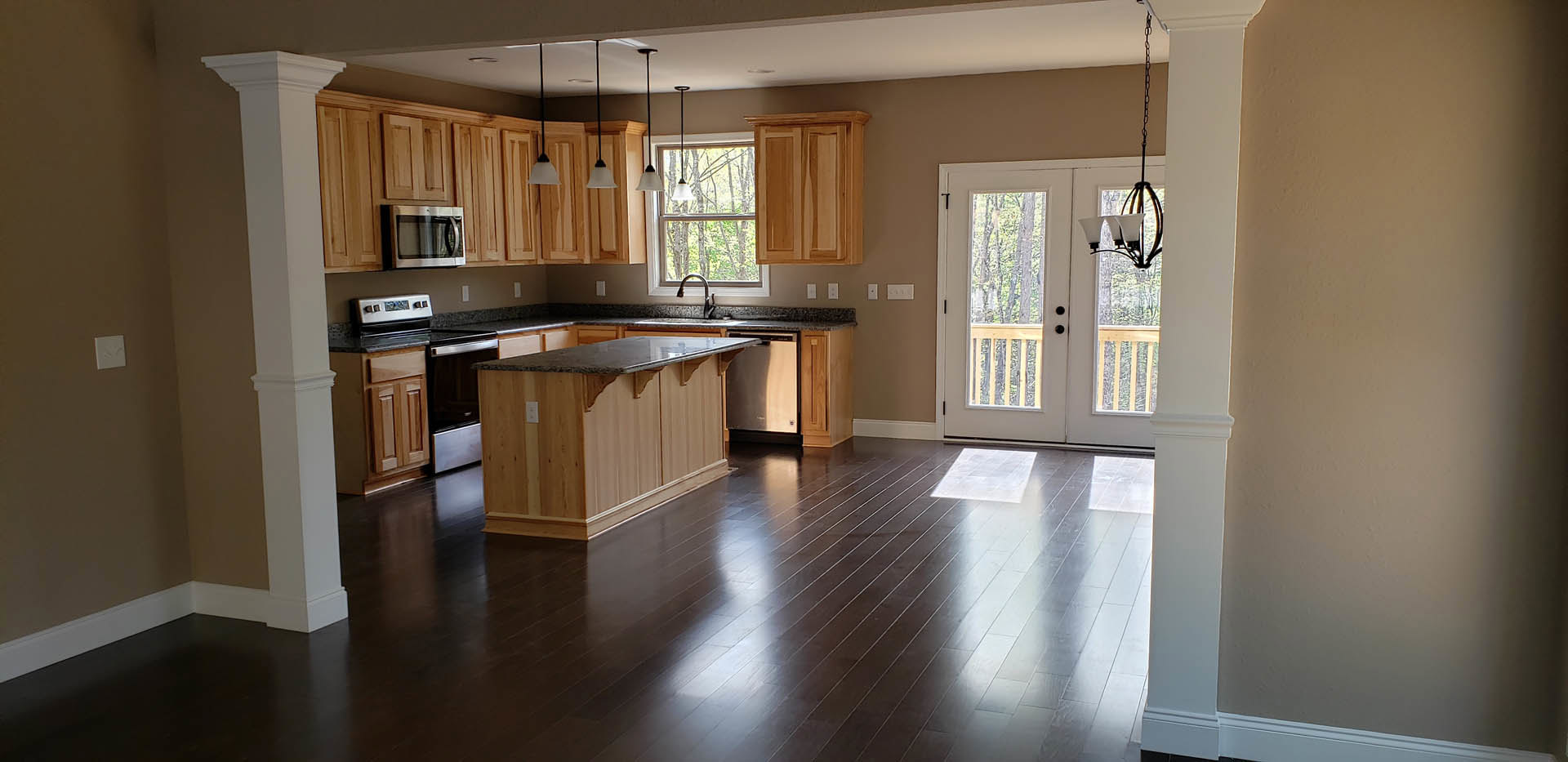 Kitchen with natural wood cabinets, matching wooden floor, granite-topped island, open microwave, close-up of white oven, double doors, and chandelier.