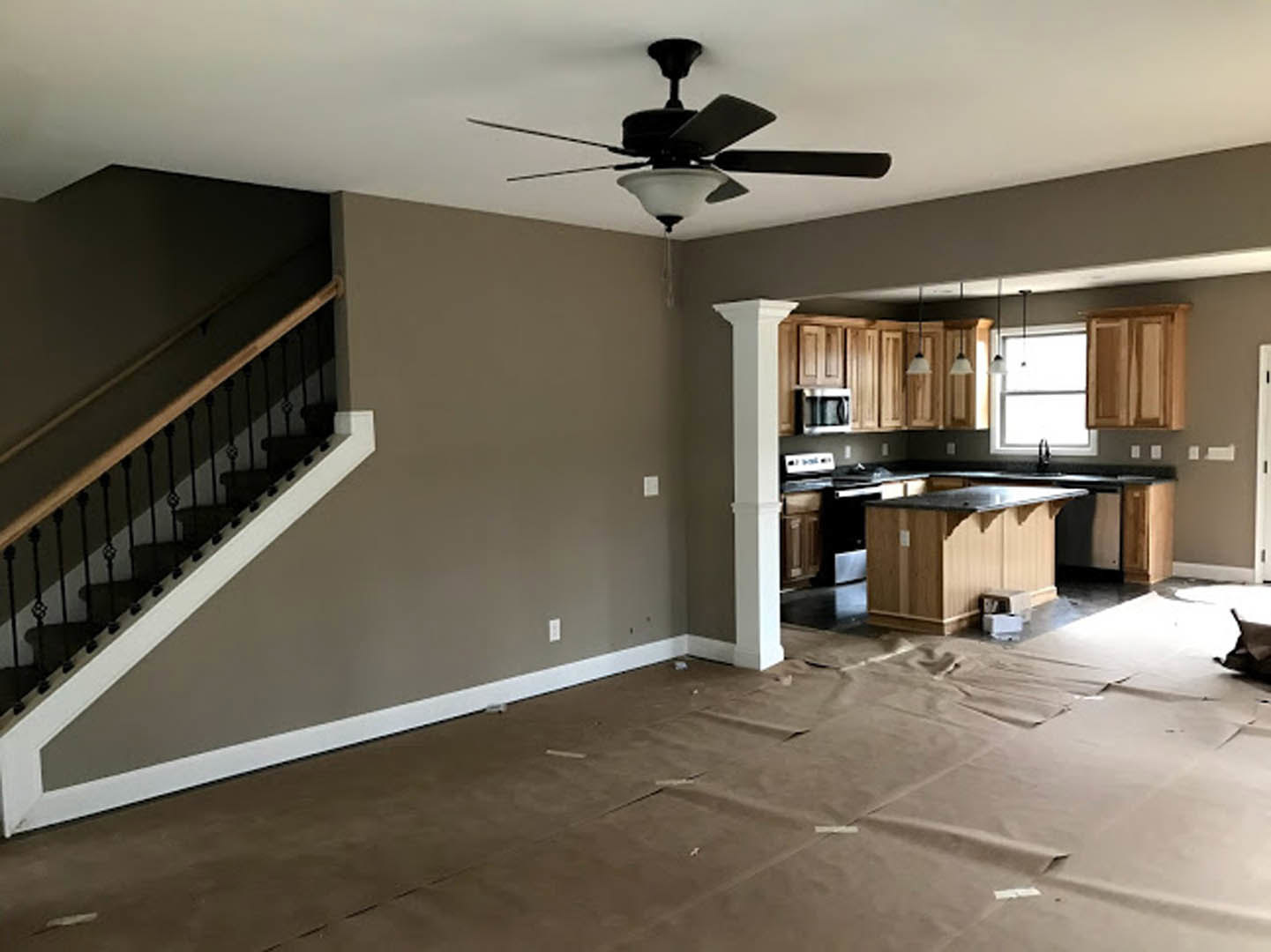 Open-concept kitchen with wooden cabinetry, black countertop, brown flooring, white trim, and ceiling fan with light fixture