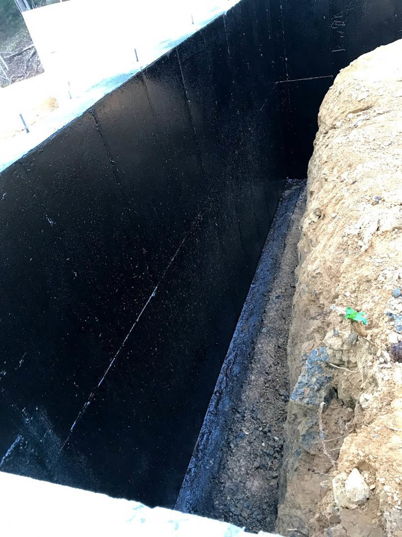 Black exterior wall with dampproofing, exposed dirt and rocks at ground level, concrete foundation visible.