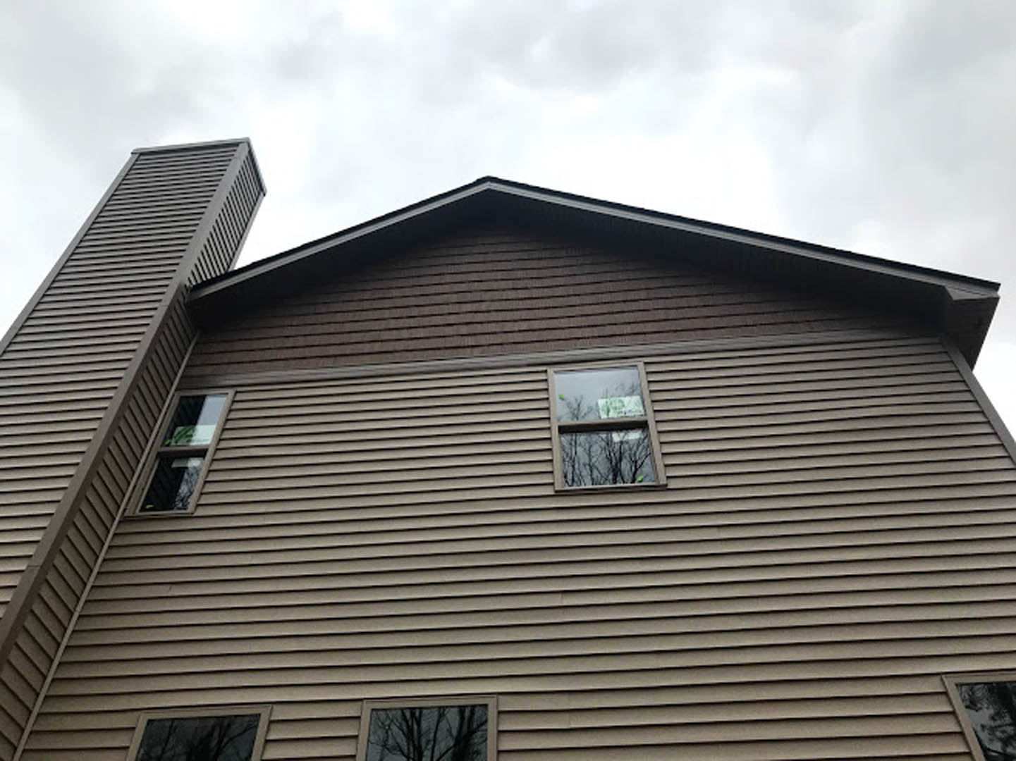 Two-story house with gray siding, brick chimney, white-trimmed windows, and tree branches against a cloudy sky