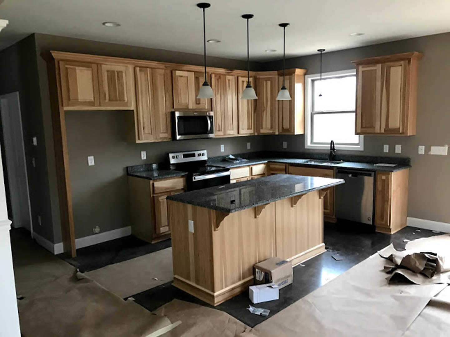 Kitchen with natural wood cabinets, stone countertop, stainless steel microwave and stove, recessed ceiling lights, window with sunlight, and a cardboard box on the floor