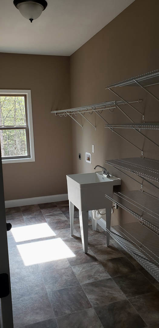 White laundry room sink with chrome faucet, gray tile floor, built-in shelves, window overlooking trees, and plaster walls
