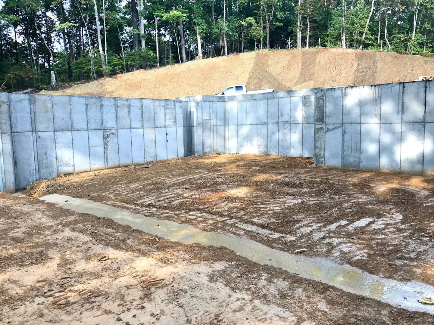 Concrete foundation wall bordered by dirt and soil, puddle of water on ground, parked truck near grassy field with trees, close-up of textured dirt surface