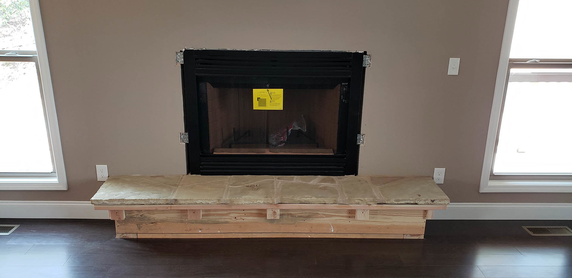 Black fireplace with stone bench, yellow caution sign, hardwood flooring, white light switch on wall, vent detail, and wood accent surfaces in a modern den.