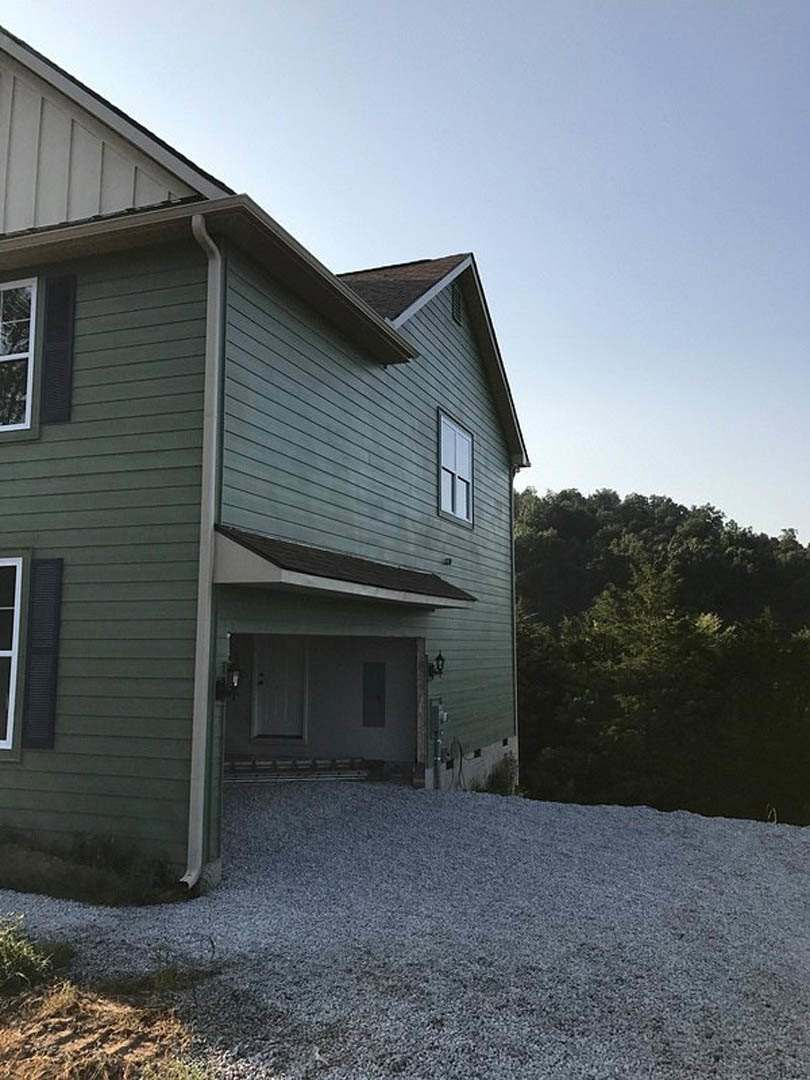 Modern home with light-colored siding, attached garage, paved driveway, white door with window, and mature trees in the background