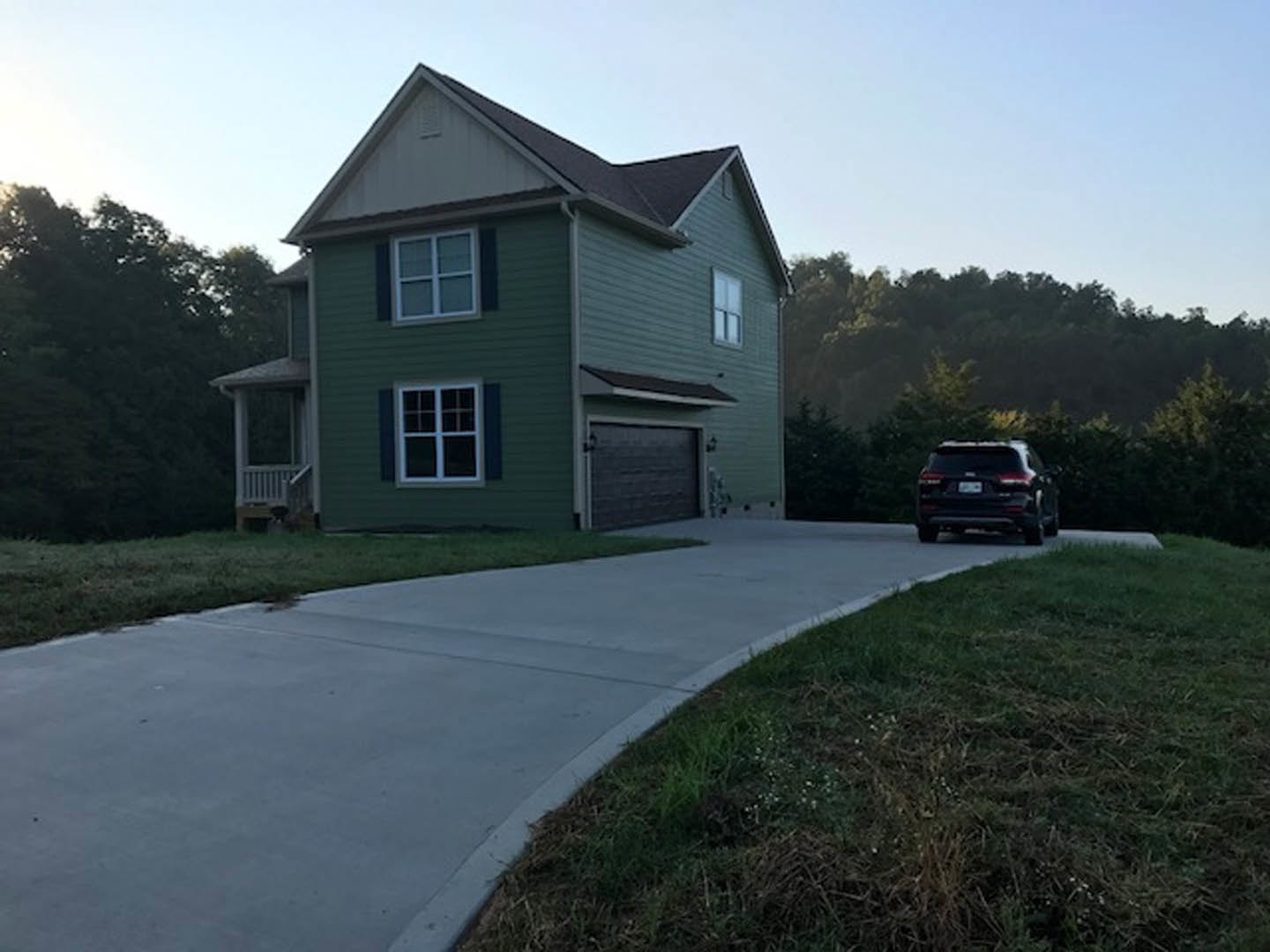 Green house with white-framed windows, attached garage with side light, driveway bordered by grass, parked car in front, leafy trees and blue sky in background